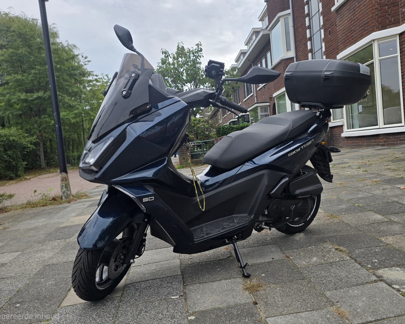Donkerblauwe Kymco Sky Town-scooter met windscherm en topkoffer geparkeerd op de stoep