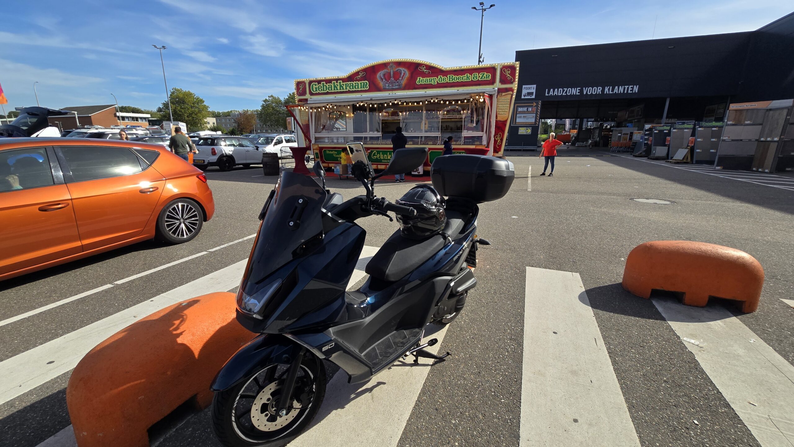 Zwarte scooter geparkeerd op een parkeerterrein met een gebakkraam op de achtergrond